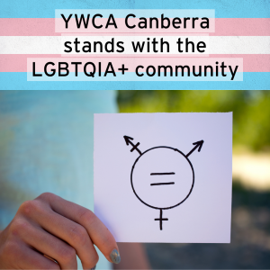 Image of a person holding a paper with the trans-inclusive gender equality symbol on it. Above, the text "YWCA Canberra stands with the LGBTQIA+ community" has a background of the trans pride flag.